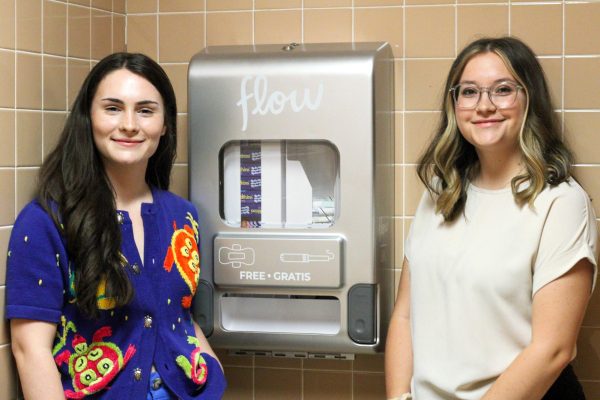 Alice Fitzgerald and Delaney Jones pose next to the first installment of the Aunt Flow Dispenser located in Ahlberg Hall on the third floor.