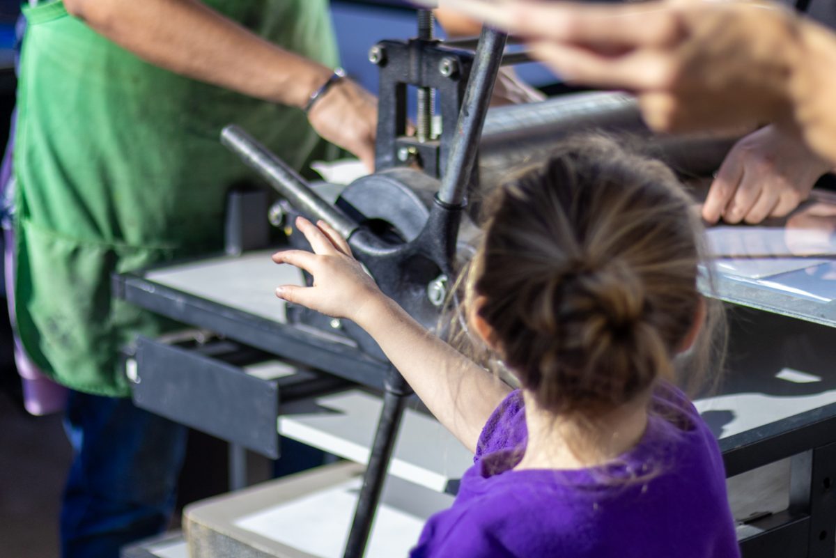 A young girl turns the printing press wheel with a member of WSU's Photography and Printmaking Guild on Nov. 23 at the Ulrich Museum's Holiday Art Cheer event.