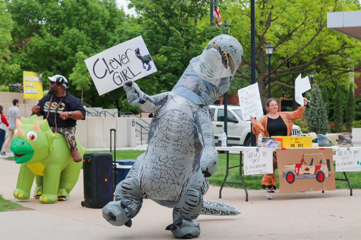 Sara Leftwich, an employee with the Shocker Career Accelerator waves a sign while dancing to motivate students before finals week. Leftwich was one of five dancing dinos who danced outside of Ablah Library on Tuesday, May 7.