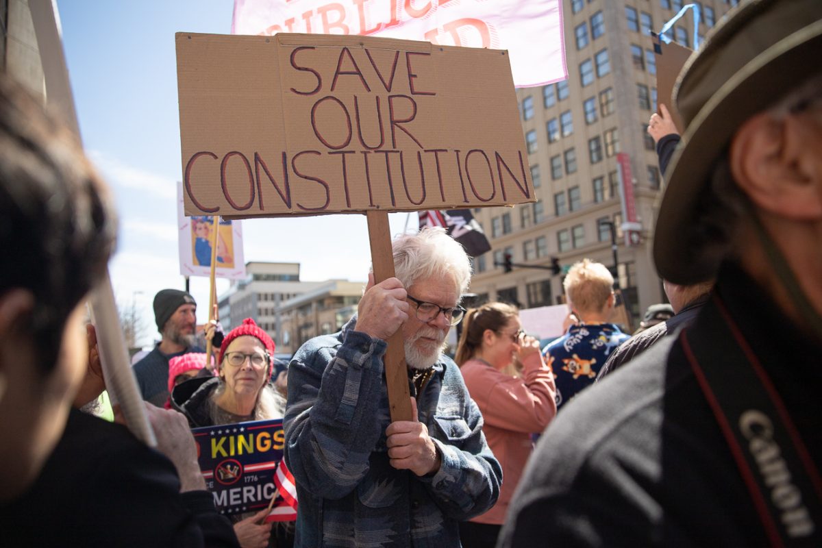 A demonstrator holds a sign reading "save our constitution" as he walks through a crowd on Saturday, March 28, 2026. Demonstrators were escorted and by police throughout the protest. (KMUW)
